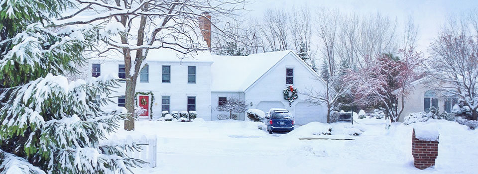 A large white house with windows across the front and a red door is in a snow-covered landscape. The house has a wreath above the garage for Christmas. There is snow on all the surrounding trees and ground.